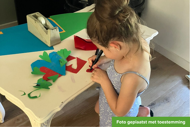 Blond meisje van 5 jaar zit op knietjes te knutselen aan tafel.