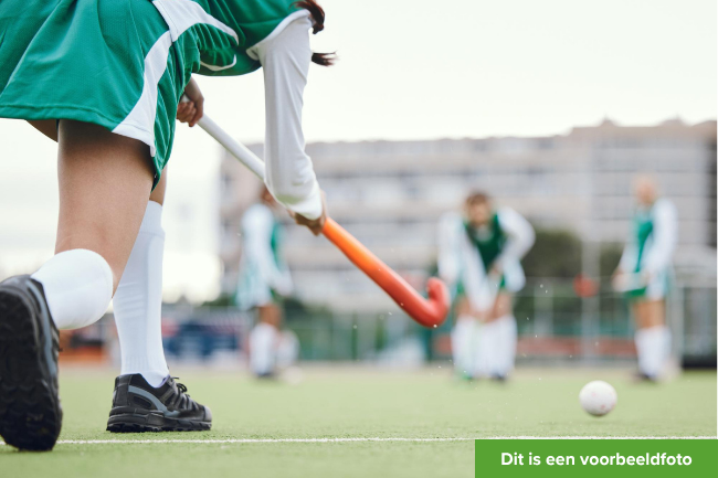 Meisje met groene sportkleding en een oranje hockeystick speelt hockey met haar team.