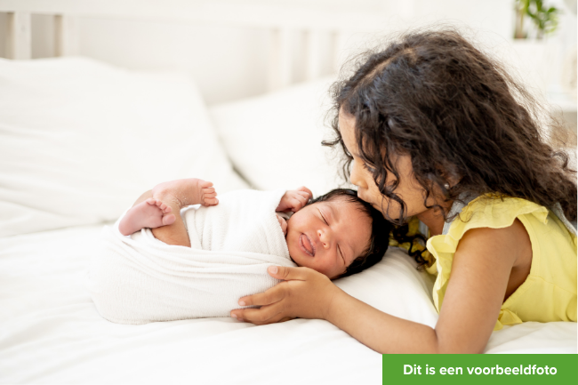 Een meisje geeft haar babybroertje of zusje een kusje op het hoofdje terwijl ze op bed liggen. Dit is een voorbeeldfoto.