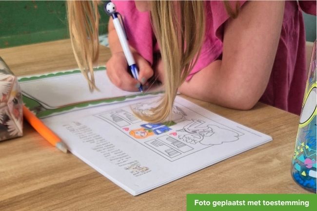 blond meisje met lang haar voorovergebogen aan tafel bezig met een vakantiedoeboek